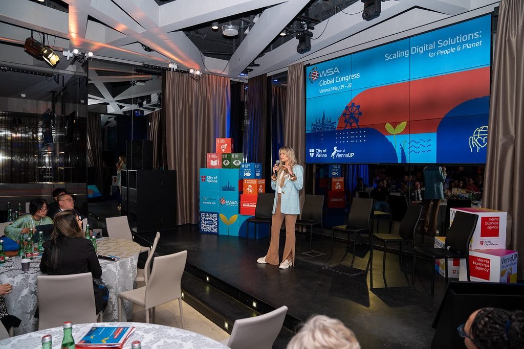 Odeta Isevičiūtė scenoje WSA Global Congress 2026, už jos kongreso ekranas, salėje prie stalų sėdi svečiai.